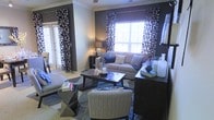 This single-floor living space showcases a thoughtfully designed interior with a cohesive navy and neutral color palette. The living area serves as the centerpiece, anchored by a deep charcoal-toned accent wall that adds depth and sophistication to the room. Bold black-and-white patterned curtains frame multiple windows, allowing natural light to filter into the space while maintaining a dramatic aesthetic. A plush, light-toned sofa is styled with navy and gray throw pillows, complementing the layered area rug beneath. A dark wood coffee table and geometric side table add visual interest and contrast against the lighter furnishings. Additional seating is provided by a woven accent chair, contributing texture and warmth to the arrangement. A media console or sideboard along one wall offers practical storage while displaying carefully chosen decorative accessories, including white ceramic vessels and sculptural accents. Access to an adjacent room or outdoor area is visible through a glass-paneled door, adding a sense of connectivity and additional natural light. The overall ambiance is polished and contemporary, blending bold design choices with comfortable, livable furnishings.