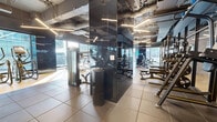 Located at 632 Newark Avenue in Jersey City, NJ, this contemporary fitness center spans a generously sized single floor, offering a well-organized and fully equipped workout facility. The interior design embraces an industrial-modern aesthetic, with exposed ceiling infrastructure, sleek linear pendant lighting, and polished black structural columns that add a striking visual element throughout the space.

The facility is divided into distinct training zones. The cardio area features a long row of elliptical machines and treadmills positioned along large windows, allowing natural light to pour in while members enjoy elevated views of the surrounding Jersey City skyline. A dedicated rowing and functional training zone benefits from expansive floor-to-ceiling glass walls on multiple sides, creating an open, light-filled environment with panoramic urban views.

The strength training section is equipped with a comprehensive selection of Matrix-brand cable machines, selectorized equipment, adjustable benches, and a full dumbbell rack. Large mirrored walls amplify the sense of space throughout. The flooring transitions between hardwood and durable rubber tile, delineating functional zones effectively. Geometric linear lighting fixtures illuminate the space uniformly, complementing the dark, high-contrast color palette of the overall design.