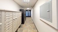 This single-floor parcel locker and mailroom area is thoughtfully designed to streamline mail and package management for residents. The elongated corridor is finished with durable, stone-look tile flooring in warm earthy tones, providing a clean and functional aesthetic throughout the space.

Along one side of the hallway, a large bank of numbered individual mailboxes in a classic cream finish offers organized mail storage for residents. Opposite and adjacent to these, a set of sleek, dark-finished Luxer One smart parcel lockers provides a modern, secure solution for package deliveries of varying sizes, complete with a digital interface panel for easy access.

The space is illuminated by recessed fluorescent ceiling panels set within a drop-tile ceiling, ensuring bright, even lighting throughout. Security is emphasized with ceiling-mounted surveillance cameras positioned along the corridor. A wall-mounted utility access panel and a mini-split HVAC unit are also present, reflecting the room's practical infrastructure.

The corridor concludes at a set of frosted glass double doors marked with unit number signage, offering a clear and secure entry point. The overall atmosphere is clean, utilitarian, and well-organized, prioritizing resident convenience and package security.