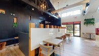 This coffee lounge is a thoughtfully designed single-floor space that seamlessly blends modern industrial aesthetics with warm, inviting elements. The layout is divided into distinct zones, each offering a unique atmosphere suited to different activities and group sizes.

The café seating area is anchored by a row of semi-private booths framed with natural wood dividers and white panels, complemented by light wood tables and contemporary chairs with gold-accented legs. Overhead, geometric brass and iron pendant chandeliers cast a warm glow against a bold black ceiling, adding a dramatic contrast to the light hardwood flooring throughout.

The open lounge area features soaring vaulted ceilings with exposed white beams and an extensive track lighting system. A whitewashed brick fireplace serves as a focal point, flanked by vibrant, large-scale mural artwork that infuses the space with energy and color. A ping pong table and eclectic seating — including a velvet swivel chair and a sleek lounge chair — make this zone ideal for casual social interaction.

Near the entrance, a curved plywood reception or service counter with bold red acrylic accents anchors the space, beside which a houndstooth-patterned decorative screen adds an artistic touch. Floor-to-ceiling windows and transom windows allow generous natural light to flood the interior, offering views of the outdoor surroundings. Trailing greenery adds organic warmth throughout the space.