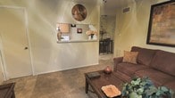 This single-floor residence offers a warm and inviting layout designed for comfortable everyday living. The living area is finished with plush wall-to-wall carpeting and centers around a wooden coffee table, complemented by a cozy upholstered sofa in rich brown tones. Decorative accents, including a woven wall piece and tasteful vases, add character to the space. A defining architectural feature is the pass-through window connecting the living area to the kitchen, topped with a convenient counter ledge that allows for easy interaction between the two spaces. The kitchen is visible through the pass-through and features light wood cabinetry. Just beyond, a dedicated dining area is finished with tile flooring and accommodates a dark dining table with matching bar-height chairs, creating a clear distinction between casual and formal eating spaces. Neutral warm tones throughout the walls and flooring lend a cohesive and welcoming ambiance to the entire home. The single-floor layout ensures seamless flow between the living, kitchen, and dining zones, making it a practical and comfortable space suited for both relaxing and entertaining.