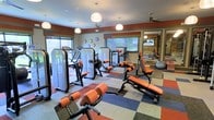 This fitness center offers a comprehensive workout environment designed to support a wide range of exercise routines. The space is outfitted with a full lineup of strength training machines featuring distinctive orange upholstery, along with adjustable benches and free weight equipment to accommodate various training styles. A large wall mirror enhances the sense of space while allowing users to monitor their form during workouts. The flooring features a colorful checkerboard pattern of red, blue, and gray padded tiles, providing both durability and cushioning underfoot. Natural light streams in through multiple windows, creating a bright and energizing atmosphere throughout. Overhead, globe pendant lights and ceiling fans are evenly distributed across the room, ensuring comfortable airflow and well-balanced illumination. The walls are accented with a bold rust-red border detail that adds a dynamic visual element to the interior. A row of clocks is mounted along one wall, and a wall-mounted screen is visible in the mirrored section of the room, offering entertainment during workouts. The overall design is functional yet visually engaging, making this fitness center a well-rounded facility suited for both strength and conditioning training.