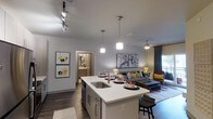 This thoughtfully designed single-floor residence showcases a bright, open-concept layout that seamlessly integrates the kitchen, dining, and living areas into one cohesive, flowing space. The kitchen serves as the heart of the home, anchored by a spacious island with a sleek white quartz countertop, an undermount sink, and seating for casual dining. Stylish pendant lighting hangs above the island, complementing a modern track lighting system overhead that ensures excellent illumination throughout the space. Cabinetry in a soft gray tone adds a contemporary touch, while stainless steel appliances contribute to the kitchen's polished, functional aesthetic. Rich dark hardwood flooring extends throughout, providing a warm contrast to the light gray walls and white ceilings. The open living area benefits from natural light streaming in through large windows dressed with teal drapery, creating a welcoming and airy ambiance. A ceiling fan enhances comfort in the living zone, which is furnished with a comfortable sofa and coordinating accent pieces. Framed artwork adorns the walls, adding character and color to the space. A hallway visible from the main area leads to a bathroom, hinting at additional private quarters beyond the open living space.