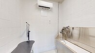 Located in Cedar Creek, TX, this dedicated pet grooming room is a compact, single-room space thoughtfully designed for professional pet care. Constructed with white-painted concrete masonry unit (CMU) block walls, the space conveys a clean, hygienic, and utilitarian character well-suited to its purpose.

The room's centerpiece is a large stainless steel grooming tub, complete with a handheld showerhead, integrated faucet, a non-slip grated floor insert, and a stainless steel splash guard. Adjacent to the tub sits a professional grooming table featuring a non-slip rubberized surface and an overhead grooming arm with restraint loops, ensuring safe and efficient grooming sessions. A standalone pet dryer is also present, adding to the room's full suite of grooming capabilities.

Climate comfort is provided by a wall-mounted mini-split air conditioning unit, ensuring a controlled environment for both pets and groomers year-round. The space features a glass-panel exterior door with a push-bar hardware, allowing natural light to filter in while providing direct exterior access — a practical feature for pet handling. The smooth, light-colored flooring completes the clean and easy-to-maintain aesthetic of this well-equipped grooming space.