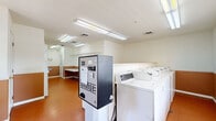 This shared laundry facility is a well-organized, single-room space designed for efficient communal use. The room features durable terracotta-toned tile flooring and two-tone walls with a warm tan wainscoting below a neutral white upper section, giving the space a clean and functional aesthetic. Overhead fluorescent lighting ensures the room is brightly illuminated throughout.

The main laundry area is equipped with a row of top-load washing machines and a stacked dryer unit, providing ample capacity for multiple users. A centrally positioned coin-change machine offers convenient access to laundry payment. Instructional signage for both washing and drying is posted along the walls for user guidance.

A dedicated folding and sorting area features a long folding table and wall-mounted wire shelving for hanging garments, making it easy for users to manage their laundry from start to finish. Multiple windows fitted with horizontal blinds allow natural light to enter the space, contributing to an open and airy feel. The room also includes clearly marked exit doors, a security camera, and a fire safety system, reflecting attention to resident safety and comfort. Overall, this laundry room is a practical and well-appointed communal amenity.