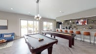 The Flats of Fishers Marketplace community clubhouse is a thoughtfully designed single-floor amenity space that combines social, recreational, and functional areas into one cohesive environment. The game room serves as a central social hub, featuring a full-size pool table and shuffleboard table beneath a stylish globe pendant light, with large sliding glass doors that flood the space with natural light and open directly to the outdoor pool area. Adjacent to the game room, a spacious community kitchen impresses with expansive granite countertops, dark cabinetry, a textured stone accent wall adorned with open shelving and greenery, pendant lighting, and a built-in beverage cooler — ideal for entertaining and gatherings. A cozy lounge area offers comfortable leather chairs with ottomans, a built-in window bench seat, and a dining nook with modern hairpin-leg furniture, all accented by herringbone-patterned wood-look flooring. A private office area enclosed by glass-paneled doors provides a quiet workspace with dedicated desks, bold accent wallcovering, and ample natural light through slatted blinds. The overall aesthetic throughout balances modern design with warm, inviting touches, creating a versatile and welcoming community space.