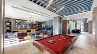 Located at 1650 NE 124th St in North Miami, this well-appointed game room delivers an energetic yet refined entertainment experience across a single open floor. The space is anchored by a striking herringbone-patterned hardwood floor that runs throughout, lending warmth and visual continuity to the entire area. Multiple pool tables with rich red felt surfaces are positioned throughout the main gaming zone, complemented by a foosball table and classic arcade cabinets, offering a diverse range of entertainment options. Large floor-to-ceiling glass windows and partitioned glass walls flood the space with abundant natural light while providing views of the surrounding outdoor areas, creating an airy, open atmosphere despite the space's intimate scale. A dedicated lounge area features a bold yellow leather banquette set against a slate-gray accent wall, surrounded by plush navy blue swivel chairs and brushed gold accent tables. The lounge wall is adorned with a curated gallery of framed sports memorabilia, including jerseys and black-and-white photography, reinforcing the sporty, upscale character of the room. Warm wood built-in shelving and dark structural columns add architectural depth, while recessed lighting and a stylish slatted ceiling detail complete the polished, contemporary aesthetic.