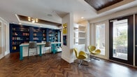 Located at 10000 Palma Linda Way in Orlando, Florida, this thoughtfully designed single-level amenity space blends contemporary style with practical functionality. The standout feature is the striking herringbone-patterned hardwood flooring that runs throughout, grounding the space with warmth and visual interest.

A dedicated library and coworking area anchors one end of the space, showcasing floor-to-ceiling navy blue built-in bookshelves filled with books and decorative objects. A teal-accented communal work table with a white surface and comfortable bar-height seating offers a functional workspace beneath a recessed tray ceiling with rich wood paneling and modern globe pendant lighting.

The open layout flows toward a bright reception or lounge area, where pairs of chartreuse swan-style chairs are artfully arranged near French doors that flood the interior with natural light and provide direct access to an outdoor terrace with views of palm trees. Open white wall shelving flanks this area, adding a clean architectural contrast to the bold color palette.

Bold, colorful abstract artwork is displayed throughout, contributing to a curated, gallery-like atmosphere. The space also features glass-paned interior French doors, adding transparency and enhancing the sense of openness. Overall, the design strikes a balance between energetic, modern aesthetics and a welcoming, community-oriented environment.