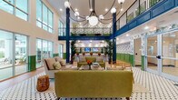 This two-story amenity space makes a striking first impression with its soaring double-height atrium, anchored by a dramatic globe chandelier and bold navy blue structural columns. The main level opens into a generously sized social lounge furnished with mid-century modern seating in olive green upholstery, arranged around a central coffee table atop a classic hexagonal tile rug. Floor-to-ceiling windows along one wall flood the space with natural light and offer direct views of an outdoor pool area, seamlessly connecting the interior to the exterior.

Throughout the space, a cohesive tropical design theme is expressed through lush monstera-leaf wallpaper, bold green wainscoting, and potted palms that reinforce the resort-like ambiance. A dedicated media lounge on the main floor features built-in wall-mounted screens, cozy bench seating, and the same distinctive botanical wallcovering, making it ideal for group viewing or casual gatherings.

The full-length catering or demonstration kitchen is outfitted with expansive white quartz countertops, stainless steel appliances, a bar sink, and ample shelving — all bathed in natural light from large windows overlooking the pool. Upstairs, a mezzanine level overlooks the atrium below, accessible via a railed balcony, and provides additional seating and gathering space with beadboard ceilings and natural wood flooring throughout.