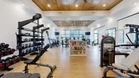 Located at 2604 Secretariat Blvd in Grove City, Ohio, this fitness center offers a thoughtfully designed, single-floor layout divided into two distinct exercise rooms. The main workout area impresses with its elevated tray ceiling clad in warm wood planking, recessed lighting, and ceiling fans that enhance the open, airy atmosphere. Light hardwood-style flooring runs throughout, contributing to the clean, modern aesthetic. This primary room is comprehensively outfitted with a row of treadmills, elliptical machines, stationary and spin bikes, cable machines, a functional trainer, adjustable benches, dumbbells, battle ropes, and a central display rack stocked with reading materials. Wall-mounted televisions and large windows with wood-trimmed blinds allow natural light to fill the space while offering outdoor views. Glass partitions connect this area to an adjacent lounge, visible through elegant chandelier lighting. The second exercise room is a bright, flexible studio space featuring soaring ceilings, expansive windows overlooking greenery, and an open floor plan ideal for stretching, yoga, or group fitness. Stability balls, dumbbells, and exercise mats are neatly organized throughout. Together, the two rooms provide a comprehensive and well-appointed environment suited for a wide range of fitness activities.