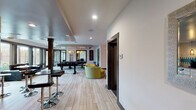 This single-floor communal amenity space is thoughtfully designed to accommodate both recreation and productivity. Light wood-look tile flooring flows consistently throughout, unifying the various functional zones within the open layout.

The game room serves as a central highlight, featuring a pool table and foosball table positioned beneath striking geometric gold pendant lights. Floor-to-ceiling windows and glass doors along one wall provide direct views and access to an outdoor pool area, filling the space with natural light.

Adjacent to the game room, a comfortable lounge area is furnished with a large sectional sofa, modular wood coffee tables, and a shag area rug. Two large-scale textured artworks anchor the walls, while an industrial-style multi-arm chandelier with exposed Edison bulbs adds a bold design statement overhead.

The kitchenette is outfitted with white cabinetry, a stainless steel under-counter refrigerator, a double sink, and a distinctive tiled backsplash featuring a teal and white geometric pattern. Amber glass pendant lights provide warm task lighting.

A dedicated conference room includes a glass-topped table surrounded by black leather and chrome chairs, set beneath a deep plum-painted ceiling with wire-cage pendant fixtures, creating a focused, professional atmosphere.