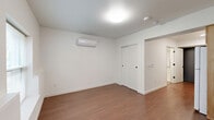This well-designed single-floor unit offers a thoughtfully organized layout that maximizes functionality across its compact footprint. The living room serves as the central gathering space, featuring warm-toned hardwood-style flooring, crisp white walls, and a wall-mounted mini-split air conditioning unit for year-round climate control. A window allows natural light to filter in, offering a view of the surrounding greenery outside. The space flows directly into the galley-style kitchen, which is outfitted with warm wood cabinetry, white laminate countertops, a stainless steel sink, a white electric range with overhead ventilation, and a full-size refrigerator — providing ample storage and functionality for everyday cooking. The bathroom is finished in clean white tones and includes an accessibility-friendly roll-in shower equipped with a fold-down bench seat and chrome grab bar, reflecting a thoughtful, inclusive design. A dedicated closet provides convenient storage. Throughout the unit, consistent hardwood-style flooring, flush ceiling fixtures, and a neutral white palette create a clean, cohesive ambiance that is both practical and welcoming.