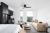 This contemporary studio presents a thoughtfully designed open-plan layout that seamlessly integrates the sleeping, living, and kitchen areas into a single, cohesive space. Light hardwood flooring runs throughout, complementing the bright white walls and ceiling, while a ceiling fan adds a practical touch overhead.

The kitchen is a standout feature, finished in sleek dark cabinetry that extends floor to ceiling, creating a bold contrast against the lighter surroundings. It is fully equipped with stainless steel appliances, including a full-sized refrigerator, range with oven, microwave, and dishwasher. Dark countertops and a stainless steel undermount sink complete the polished, modern aesthetic.

The living area is positioned toward the large floor-to-ceiling windows, which flood the space with natural light and offer sweeping city views — a defining characteristic of this elevated unit. A seating arrangement with a sofa, accent chairs, and a striped area rug defines the lounge zone, while the sleeping area is situated adjacent to the kitchen block, providing a natural sense of separation within the open layout.

Overall, the space delivers a modern, urban aesthetic with clean lines, high-quality finishes, and an abundance of natural light.