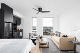 This contemporary studio presents a thoughtfully designed open-plan layout that seamlessly integrates the sleeping, living, and kitchen areas into a single, cohesive space. Light hardwood flooring runs throughout, complementing the bright white walls and ceiling, while a ceiling fan adds a practical touch overhead.

The kitchen is a standout feature, finished in sleek dark cabinetry that extends floor to ceiling, creating a bold contrast against the lighter surroundings. It is fully equipped with stainless steel appliances, including a full-sized refrigerator, range with oven, microwave, and dishwasher. Dark countertops and a stainless steel undermount sink complete the polished, modern aesthetic.

The living area is positioned toward the large floor-to-ceiling windows, which flood the space with natural light and offer sweeping city views — a defining characteristic of this elevated unit. A seating arrangement with a sofa, accent chairs, and a striped area rug defines the lounge zone, while the sleeping area is situated adjacent to the kitchen block, providing a natural sense of separation within the open layout.

Overall, the space delivers a modern, urban aesthetic with clean lines, high-quality finishes, and an abundance of natural light.