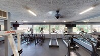 This fitness center is a dedicated, single-floor exercise facility designed to support a comprehensive workout experience. The main workout area is generously sized, housing a strong selection of cardio equipment including multiple treadmills, elliptical trainers, and a stair climber, all arranged to maximize use of the available floor space. A multi-station strength training machine and weight bench provide options for resistance training alongside the cardio offerings. A full-length wall mirror spans one side of the room, enhancing the sense of openness and allowing users to monitor their form during exercise. Large windows line the perimeter, flooding the space with natural light and offering views of the surrounding outdoor greenery and pool area. The floors are finished with durable rubber gym flooring, and a ceiling fan provides additional airflow throughout the space. Recessed fluorescent lighting and a wall-mounted television add to the functional amenities. An adjoining entryway area features blue-gray walls, a water dispenser, and access to additional rooms including restrooms or changing areas, completing the facility's practical layout. The textured ceilings and neutral color palette give the space a clean, focused atmosphere suited for fitness use.