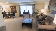 This single-floor space centers around a generously sized living area finished with plush wall-to-wall carpeting in a neutral tone, creating a warm and inviting atmosphere. A large picture window dressed with white blinds and floor-length curtains serves as the focal point of the room, allowing abundant natural light to fill the space. The window is framed by a sleek dark wood console table, accessorized with decorative objects that add a refined touch.

The seating area features a substantial gray upholstered sofa adorned with patterned throw pillows, paired with a tufted dark leather ottoman that doubles as a coffee table. A tall floor lamp anchors one corner, complementing the ambient lighting throughout. Lush potted plants are thoughtfully placed to introduce organic texture and color contrast against the neutral walls.

Adjacent to the living area, a dining space is partially visible, furnished with dark wood chairs and framed wall art, maintaining a cohesive, understated aesthetic. The overall ambiance is calm and well-coordinated, blending functional layout with classic, contemporary décor.