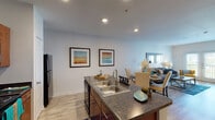 This single-floor residence is designed around an open-concept layout that seamlessly connects the kitchen, dining, and living areas into one cohesive space. The kitchen serves as a central hub, anchored by a peninsula with dark granite-style countertops and a double stainless steel sink. Rich wood-toned cabinetry lines the walls, providing generous storage, while black appliances — including a refrigerator, electric range, and built-in microwave — offer a bold contrast. Wood-look flooring flows throughout the main living areas, adding warmth and continuity.

The dining area sits adjacent to the kitchen peninsula, transitioning naturally into the living room, which benefits from natural light through glass patio doors that open to a small balcony with outdoor views. The living space is bright and versatile, complemented by neutral wall tones and recessed lighting.

The bedroom is a comfortable retreat featuring a large window that draws in ample natural light and provides exterior views. Warm, neutral tones and carpeted flooring contribute to a relaxed atmosphere.

The bathroom is finished with wood-look flooring, a vanity with warm countertops and wood cabinetry, and a full tub-shower combination with tile surround, maintaining the consistent, cohesive design found throughout the unit.