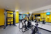 Located at 13223 Champions Centre Dr in Houston, TX, this fitness center delivers a fully outfitted workout experience within a compact, single-floor space. The interior is immediately striking, with bold yellow, orange, and red accent walls serving as the backdrop for large-scale athletic murals that energize the environment and reinforce the space's purpose. A particularly eye-catching mural depicting dynamic figures in motion spans one full wall, adding an artistic, motivational character to the room.

The facility is equipped with a well-rounded selection of exercise equipment. Cardio options include elliptical trainers, a stationary spin bike, and a treadmill. Strength and functional training is supported by a Matrix cable and functional trainer system, a weight bench, a dumbbell rack, plyo boxes, medicine balls, resistance bands, and stability balls. Wall-mounted storage keeps accessories organized and accessible.

Large mirrors line one wall, visually expanding the space and providing users with form-checking capability during workouts. Recessed linear lighting ensures the room is evenly and brightly lit throughout. A wall-mounted flat-screen TV adds a convenient entertainment option. The flooring consists of durable, cushioned rubber-style material appropriate for high-impact activity. The overall ambiance is high-energy, purposeful, and thoughtfully designed to maximize utility within its footprint.