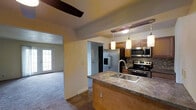 This single-floor residence is designed with an open-concept layout that seamlessly connects the kitchen and living areas. The kitchen serves as the centerpiece of the space, featuring a peninsula with a double stainless steel sink and granite-style countertops, offering both functionality and a natural gathering point. Warm wood cabinetry lines the walls, complemented by a decorative tile backsplash in a mosaic pattern that adds texture and visual interest. A suite of stainless steel appliances — including a French door refrigerator with an in-door ice and water dispenser, a microwave, and an electric range — provides a cohesive, modern finish. Three cylindrical pendant lights hang above the peninsula, casting warm, ambient light over the workspace. The adjoining living area features plush carpeting and is bathed in natural light streaming through a glass-paneled exterior door, creating a bright and welcoming atmosphere. A ceiling fan is positioned overhead, adding both comfort and style. The overall design blends warm tones, practical finishes, and an efficient use of space, making it an inviting and functional home environment.