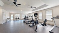 Located at 200 Governors Ct in Cartersville, GA, this fitness center offers a spacious, single-floor layout designed to accommodate a comprehensive workout experience. The open floor plan is finished with durable, textured rubber flooring throughout, providing a safe and functional surface suited for a range of exercise activities.

The facility is well-lit by multiple large windows fitted with slatted blinds, allowing abundant natural light to fill the space while maintaining privacy. Two ceiling fans are mounted overhead — including one positioned beneath a raised tray ceiling feature — promoting consistent airflow and comfort during workouts. Recessed lighting supplements the natural light for well-distributed illumination throughout.

The cardio section is equipped with treadmills, elliptical machines, and a BodyCraft rowing machine, while a wall-mounted flat-screen TV provides entertainment during exercise. Strength training options include a multi-function cable machine and a TAG weight bench with free weights, positioned in a quieter corner of the room alongside a water fountain for convenience. A dedicated seating area near the entrance features wooden chairs, offering a welcoming spot to rest.

Glass-panel doors provide direct access to the exterior, with views of the surrounding outdoor area visible through the windows. The clean, neutral-toned walls and thoughtful layout create a bright, functional, and comfortable environment for fitness enthusiasts.