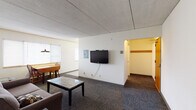 This single-floor unit offers a straightforward, functional layout designed to maximize use of space across its main living areas. The combined living and dining zone serves as the heart of the unit, featuring patterned carpeting, neutral white walls, and a textured ceiling. A wall-mounted flat-screen television anchors one side of the living area, while a gray tufted sofa and a sleek coffee table provide comfortable seating. A wooden dining table with chairs sits near a window fitted with white horizontal blinds, allowing natural light to filter into the space.

The kitchen is laid out in an efficient galley configuration, offering ample white cabinetry with warm wood-tone trim and generous counter space. White appliances, including an electric range, microwave, dishwasher, and full-size refrigerator, provide complete cooking functionality. Recessed under-cabinet lighting brightens the workspace.

The bedroom is a quiet, carpeted room with a window and baseboard heating, offering a simple and restful environment. The bathroom features a white vanity with under-sink storage, a large mirror, and light, neutral flooring. A coat hook near the bathroom entry adds a practical touch. Overall, the unit presents a clean, utilitarian aesthetic suited for comfortable everyday living.