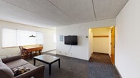 This single-floor unit offers a straightforward, functional layout designed to maximize use of space across its main living areas. The combined living and dining zone serves as the heart of the unit, featuring patterned carpeting, neutral white walls, and a textured ceiling. A wall-mounted flat-screen television anchors one side of the living area, while a gray tufted sofa and a sleek coffee table provide comfortable seating. A wooden dining table with chairs sits near a window fitted with white horizontal blinds, allowing natural light to filter into the space.

The kitchen is laid out in an efficient galley configuration, offering ample white cabinetry with warm wood-tone trim and generous counter space. White appliances, including an electric range, microwave, dishwasher, and full-size refrigerator, provide complete cooking functionality. Recessed under-cabinet lighting brightens the workspace.

The bedroom is a quiet, carpeted room with a window and baseboard heating, offering a simple and restful environment. The bathroom features a white vanity with under-sink storage, a large mirror, and light, neutral flooring. A coat hook near the bathroom entry adds a practical touch. Overall, the unit presents a clean, utilitarian aesthetic suited for comfortable everyday living.
