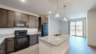 This one-bedroom, one-bathroom unit in Denton, TX offers a thoughtfully designed, single-floor layout that maximizes every square foot of its compact footprint. The open-concept kitchen and living area serves as the heart of the home, anchored by a central island with a granite countertop and an integrated stainless steel sink, illuminated by stylish pendant lighting. Rich, dark-toned cabinetry lines the kitchen walls, providing generous storage, while matching granite countertops and black appliances — including a refrigerator, range, and microwave — complete the cohesive look. Light wood-style flooring runs throughout the main living area, adding warmth and continuity. The bedroom is a comfortable, carpeted retreat featuring a ceiling fan, a window that brings in natural light, and access to a dedicated closet. The bathroom is cleanly appointed with a tiled walk-in shower, a granite-topped vanity with dark cabinetry, and wood-style flooring. Additional practical spaces include a utility room, laundry area, and ample closet storage. A sliding glass door off the living area provides access to a balcony, offering a touch of outdoor connection. The overall design is modern and consistent, with neutral tones and quality finishes throughout.