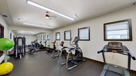 This dedicated fitness center offers a comprehensive workout environment designed to support a wide range of exercise routines. The space is laid out across a single floor with a practical, elongated floor plan that maximizes equipment placement and ease of movement.

The cardio section is well-appointed with multiple treadmills, elliptical trainers, upright stationary bikes, and a recumbent bike, providing ample options for aerobic exercise. A multi-functional strength training station anchors another area of the room, suitable for a variety of resistance exercises. The flooring throughout is durable rubber, ideal for fitness use and easy maintenance.

Large wall-mounted mirrors run along one side of the room, visually expanding the space and allowing users to monitor their form during workouts. Rows of dark-trimmed windows line the walls, inviting natural light and offering views of the surrounding exterior. Additional illumination is provided by overhead fluorescent lighting and a ceiling fan that promotes air circulation.

A set of glass-paneled doors offers direct access to an outdoor area, enhancing ventilation and connectivity to the exterior. The walls are finished in a clean, neutral tone, contributing to a bright and motivating atmosphere. Overall, this fitness center is thoughtfully organized to deliver a fully functional and comfortable exercise experience.
