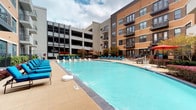 This expansive outdoor amenity area is centered around a large, sparkling swimming pool with clearly marked depth indicators and a pool volleyball net, making it ideal for both relaxation and recreation. The pool is framed by a generously sized deck finished in natural stone and brick pavers, offering ample space for lounging and socializing. Vibrant blue chaise lounges and reclining chairs are arranged poolside, complemented by shaded seating areas featuring bold orange and red canopy sails and umbrellas. A dedicated outdoor grilling station with a stainless steel built-in grill and granite countertop provides a fully equipped space for outdoor dining and entertaining. A raised composite wood deck along the building perimeter offers an additional lounge zone furnished with wicker chairs and lush potted greenery. The area is surrounded by mature ornamental trees and well-maintained landscaping, lending a private, courtyard-like atmosphere. The multi-story brick and contemporary facade of the building encloses the space on multiple sides, creating a sense of enclosure and community while still allowing for open sky above.