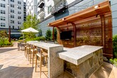 This thoughtfully designed outdoor courtyard offers a versatile and inviting gathering space set within a modern urban building complex. The ground-level patio features light-toned stone pavers that extend across the open area, creating a clean, cohesive foundation throughout. Multiple distinct zones are arranged across the courtyard to accommodate a variety of activities and group sizes.

A large dining area anchors one section of the space, furnished with a generous rectangular wooden table surrounded by cushioned chairs and shaded by a substantial white market umbrella. Overhead string lights are strung across the courtyard, adding warmth during evening hours. Adjacent to the dining area, a wood-clad pavilion with warm horizontal slatted panels houses a wall-mounted flat-screen television, making it well-suited for outdoor entertainment.

A long granite-topped bar counter runs parallel to the pavilion structure, lined with wooden barstools and framed beneath a striking slatted wood pergola. The bar area also includes built-in grill equipment, enhancing the space's outdoor kitchen functionality.

Throughout the courtyard, lush landscaped beds feature trees, ornamental grasses, and shrubs, softening the surrounding architecture. A lounge seating area with cushioned chairs and a sofa adds a relaxed social zone. Outdoor floor lamps and warm wood cladding on surrounding walls complete the refined, resort-like ambiance.