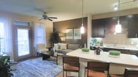 This single-floor residence showcases a well-appointed open-concept floor plan that integrates the living, dining, and kitchen spaces into one cohesive and functional environment. The living area is anchored by a bold dark accent wall, which provides a striking contrast against the neutral surrounding tones, and is complemented by a ceiling fan that enhances comfort and air circulation. Large windows dressed with bright white curtains invite natural light into the space, creating a warm and welcoming ambiance. The kitchen is finished with rich dark cabinetry, a classic subway tile backsplash, and a light-toned granite or stone countertop that extends into a curved breakfast bar with seating — ideal for casual dining or entertaining. A modern pendant light hangs above the bar, adding a refined focal point to the area. The open layout allows for a smooth visual and physical flow between the kitchen and the living room, making the space feel expansive and connected. Thoughtful design details throughout, including the layered lighting, decorative greenery, and coordinated furnishings, contribute to an atmosphere that is both stylish and livable.