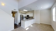 This two-story space showcases a thoughtfully designed open-concept layout where the kitchen and living areas flow naturally into one another. The kitchen serves as a central focal point, featuring a peninsula with a grey quartz-style countertop that provides both workspace and a casual dining option. White shaker-style cabinetry lines the walls, complemented by stainless steel appliances including a refrigerator, microwave, and range. The grey countertops offer a clean contrast against the bright white cabinetry, creating a modern, cohesive aesthetic. Dark hardwood-style flooring anchors the kitchen area, transitioning smoothly into plush carpeting in the adjacent living space, defining each zone without the need for walls. Neutral beige tones throughout the walls contribute to a bright, welcoming atmosphere. Recessed lighting and a ceiling-mounted fixture provide ample illumination. Notably tall ceilings amplify the sense of openness, further enhanced by the two-floor design. A staircase is visible, connecting the main living level to the upper floor, adding both functional connectivity and architectural interest to the overall space.