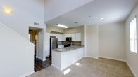 This two-story space showcases a thoughtfully designed open-concept layout where the kitchen and living areas flow naturally into one another. The kitchen serves as a central focal point, featuring a peninsula with a grey quartz-style countertop that provides both workspace and a casual dining option. White shaker-style cabinetry lines the walls, complemented by stainless steel appliances including a refrigerator, microwave, and range. The grey countertops offer a clean contrast against the bright white cabinetry, creating a modern, cohesive aesthetic. Dark hardwood-style flooring anchors the kitchen area, transitioning smoothly into plush carpeting in the adjacent living space, defining each zone without the need for walls. Neutral beige tones throughout the walls contribute to a bright, welcoming atmosphere. Recessed lighting and a ceiling-mounted fixture provide ample illumination. Notably tall ceilings amplify the sense of openness, further enhanced by the two-floor design. A staircase is visible, connecting the main living level to the upper floor, adding both functional connectivity and architectural interest to the overall space.