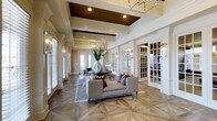 Located at 20 Ashton Village Dr in Westerville, Ohio, this sophisticated single-floor space exudes a timeless, upscale aesthetic through its carefully considered architectural design and premium finishes. The main living area immediately captivates with its dramatic coffered ceiling, accented by rich dark wood insets and recessed lighting that draw the eye upward and lend a sense of generous height to the space. An elegant geometric parquet hardwood floor serves as a stunning foundation throughout, adding warmth and visual interest underfoot. Crisp white crown molding, decorative columns, and detailed millwork frame the space with classic refinement. Large windows fitted with plantation shutters line the walls, inviting an abundance of natural light while maintaining a sense of privacy. A modern geometric chandelier anchors the ceiling, complementing the sophisticated lighting fixtures adorning the walls. French doors with glass pane detailing lead to adjoining areas, enhancing the open and airy flow of the layout. Neutral, earthy tones across the walls and furnishings create a warm and welcoming ambiance, making the space both visually impressive and functionally inviting. Overall, the design strikes a harmonious balance between classical elegance and contemporary comfort.