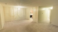 This single-floor space offers a clean and functional layout with a welcoming living area as its centerpiece. The room is finished with soft, neutral-toned carpeting that extends throughout, creating a comfortable and cohesive feel underfoot. A chandelier-style ceiling light fixture provides warm, ambient illumination, complementing the light-colored walls that lend a bright and airy atmosphere to the space.

An open doorway connects the living area to the kitchen, offering a practical flow between the two spaces. The kitchen features light wood cabinetry that adds a warm, natural tone, along with standard appliances including a range and overhead ventilation. The kitchen is finished with hard flooring, providing a functional contrast to the carpeted living area.

The walls throughout are smooth and neutral, offering a versatile backdrop suitable for a variety of furnishing styles. The textured ceiling adds a subtle design element consistent with the overall understated aesthetic of the space. The layout is straightforward and efficient, making good use of the available square footage while maintaining an open and uncluttered feel. Overall, the space presents a tidy, move-in-ready environment with all the essential elements for comfortable everyday living.