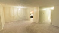 This single-floor space offers a clean and functional layout with a welcoming living area as its centerpiece. The room is finished with soft, neutral-toned carpeting that extends throughout, creating a comfortable and cohesive feel underfoot. A chandelier-style ceiling light fixture provides warm, ambient illumination, complementing the light-colored walls that lend a bright and airy atmosphere to the space.

An open doorway connects the living area to the kitchen, offering a practical flow between the two spaces. The kitchen features light wood cabinetry that adds a warm, natural tone, along with standard appliances including a range and overhead ventilation. The kitchen is finished with hard flooring, providing a functional contrast to the carpeted living area.

The walls throughout are smooth and neutral, offering a versatile backdrop suitable for a variety of furnishing styles. The textured ceiling adds a subtle design element consistent with the overall understated aesthetic of the space. The layout is straightforward and efficient, making good use of the available square footage while maintaining an open and uncluttered feel. Overall, the space presents a tidy, move-in-ready environment with all the essential elements for comfortable everyday living.