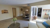 This single-floor residence features an open-concept layout that seamlessly connects the living and dining areas, creating a cohesive and functional flow throughout the space. The living area is finished with wall-to-wall carpeting in a neutral beige tone, complementing the warm, light-colored walls that give the interior a bright and welcoming ambiance. A textured ceiling adds subtle visual interest overhead.

The dining area is defined by a dark round table surrounded by upholstered chairs, illuminated by a chandelier that serves as a focal point. Decorative wall accents, including a sunburst mirror and a clock, contribute to the curated aesthetic of the space.

A structural column subtly separates the living and dining zones while maintaining visual openness. Natural light enters through a glass door that leads to an outdoor area, where woven storage pieces and a glimpse of exterior greenery are visible.

A hallway extends toward additional rooms, including doors that suggest bedroom and closet access, providing a sense of depth and privacy within the layout. Overall, the space presents a comfortable, well-connected interior with a consistent neutral palette throughout.