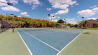 This outdoor recreational facility centers around a set of well-maintained tennis courts finished in a classic blue and green hard-court surface, clearly marked with crisp white lines. The courts are equipped with regulation nets and are supported by tall overhead flood lighting structures, making the facility suitable for both daytime and evening play. The open-air setting is framed by a dense perimeter of mature trees, including palms, which provide a natural green backdrop and a degree of shade and wind buffering. The surrounding landscape suggests a warm, subtropical climate with expansive open skies. Adjacent to the courts sits a modest single-story structure that appears to serve as a clubhouse or support facility, likely housing restrooms, storage, or administrative space. The grounds are open and spacious, allowing for comfortable movement between courts. The combination of quality court surfacing, lighting infrastructure, and supporting amenities makes this a functional and well-equipped venue suited for recreational and competitive tennis alike.