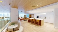 Located at 299 Duffield Street in the heart of Downtown Brooklyn, this amenity floor offers a well-appointed, single-level space designed for comfort, productivity, and wellness. The open lounge and social area immediately impresses with its warm wood-paneled ceiling, light oak flooring, and an abundance of natural light streaming through large windows. A central island with bar seating anchors the space alongside a white tile backsplash, a built-in coffee station, and soft under-cabinet lighting, complemented by communal dining tables and cozy lounge seating. A library area provides a quieter retreat, featuring shelving and a relaxed, focused atmosphere. The two gym spaces are comprehensively outfitted with cardio machines including TRUE treadmills, ellipticals, and stationary bikes, as well as a Hoist functional trainer, dumbbells, medicine balls, and a flat-screen TV — all set on durable rubberized flooring with natural light from street-facing windows. A sleek, fully accessible bathroom finished in crisp white tile with stainless steel grab bars rounds out the floor. The overall design palette of warm wood, white surfaces, and neutral tones creates a bright, modern, and welcoming environment throughout.