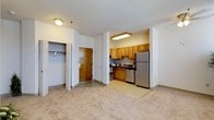 This single-floor residential unit offers a practical and well-organized layout designed for comfortable everyday living. Upon entry, a coat closet with a hanging rod provides convenient storage, positioned alongside the main entry door. The open-concept design allows the living area and kitchen to flow seamlessly together, creating a sense of spaciousness throughout.

The kitchen is nestled within a defined alcove and is equipped with warm honey-toned wood cabinetry that extends to the upper walls, offering generous storage. Stainless steel appliances, including a full-sized refrigerator and a built-in microwave above the range, lend the kitchen a modern, functional aesthetic. A double sink and additional counter space complete the cooking area, which features tile flooring that transitions smoothly into the carpeted living space.

The living area is finished with neutral, light-toned carpeting that contributes to a clean and inviting ambiance. Flush ceiling light fixtures provide consistent illumination throughout the unit. The overall color palette — soft whites and warm beiges — creates a bright and welcoming atmosphere. The layout is efficient and thoughtfully designed, making the most of the available space while maintaining a comfortable and livable environment.