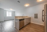 This thoughtfully designed single-floor unit offers a clean, contemporary aesthetic throughout. The open-concept kitchen flows seamlessly into the living area, anchored by a peninsula with a light-toned countertop that doubles as a breakfast bar. The kitchen is equipped with a full suite of stainless steel appliances, including a refrigerator, range, microwave, and dishwasher, complemented by sleek gray cabinetry and a stainless steel undermount sink. Warm wood-look flooring runs throughout the main living areas, adding a welcoming touch to the otherwise crisp, neutral palette of white and light gray walls. The bathroom features large-format tile flooring in a warm beige tone, offering a clean and classic look, with a full shower and toilet included. The bedroom is a standout space, bathed in natural light through tall, multi-pane windows that frame sweeping views of the surrounding cityscape and greenery. The open layout, flush-mount ceiling lighting, and neutral finishes throughout create a bright, airy ambiance that is both functional and inviting, making this a well-rounded living space suited to a variety of lifestyles.