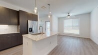This single-floor residence showcases a contemporary open-concept design that seamlessly connects the kitchen and living areas. The kitchen is a standout feature, anchored by a generous island topped with light granite countertops and equipped with an undermount sink and brushed gold pull-down faucet. Sleek dark-toned flat-front cabinetry extends to the ceiling, offering substantial storage, while a classic white subway tile backsplash provides a crisp contrast. Stainless steel appliances, including a French door refrigerator with ice and water dispenser, electric range, and built-in microwave, complete the well-appointed kitchen. Glass pendant lights hang above the island, adding a refined touch. Light wood-look flooring flows throughout the main living spaces, enhancing the bright, airy atmosphere created by large windows fitted with horizontal blinds. A ceiling fan is centrally positioned in the living area, which also benefits from direct access to an outdoor balcony. The bedroom is carpeted and features multiple windows with outdoor views and a ceiling fan. A spacious walk-in closet with wire shelving provides ample storage. The bathroom includes light wood flooring, a tiled walk-in shower with gold-accented glass doors, and matching dark cabinetry consistent with the kitchen's design language.