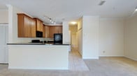 This single-floor unit features two bedrooms and one bathroom within a thoughtfully designed layout. The kitchen is a standout area, outfitted with warm honey-toned wood cabinetry offering generous storage, dark granite-style countertops, and black appliances including a refrigerator, microwave, and additional cooking appliances. A peninsula with a built-in sink extends into the space, providing additional prep area and serving as a natural divider between the kitchen and the adjoining living area. Track lighting along the kitchen ceiling ensures strong task illumination throughout. The flooring transitions from light ceramic tile in the kitchen to plush carpeting in the living areas, adding warmth underfoot. Crown molding runs along the ceiling, lending a refined finish to the interiors. A hallway visible from the main living space leads toward the bedrooms and bathroom, with closet doors suggesting ample storage. The overall palette is neutral and bright, with white walls and trim creating a clean, open atmosphere throughout the unit.