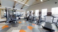This single-floor fitness center is thoughtfully designed to support a comprehensive workout experience. The space is defined by striking vaulted ceilings with exposed wooden beams, lending an open and airy architectural character that sets it apart from a typical gym environment. Linear pendant lighting runs along the ceiling, ensuring the space is well-lit throughout.

Large windows line the walls, inviting an abundance of natural light and providing views of the surrounding exterior, creating an energizing atmosphere for exercise. The flooring is a standout design feature, composed of a bold geometric pattern in shades of orange, gray, and neutral tones that adds a modern, dynamic feel to the space.

The fitness center is well-appointed with a range of cardio equipment, including elliptical trainers, stair climbers, and stationary bikes, all positioned to take advantage of the natural light from the windows. A wall-length mirror on one side enhances the sense of space while supporting strength training and form checks. A multi-functional cable and weight machine provides options for a full-body strength workout.

Overall, the space combines functional fitness amenities with intentional architectural and interior design details, resulting in a motivating and visually appealing environment for users of all fitness levels.