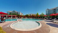 Located at 4883 Riley Rd in Bethlehem, Pennsylvania, this outdoor amenity area centers around a generously sized swimming pool with clear, blue-tinted water and tiled lane markings. The pool is complemented by a shallow wading area, providing options for both lap swimming and leisurely relaxation.

Surrounding the pool is a spacious, paved sundeck lined with rows of cushioned lounge chairs and side tables, all shaded by a generous collection of bold red patio umbrellas. The uniform arrangement of the seating creates a resort-style atmosphere designed for comfort and convenience. A pergola structure at one end of the deck adds architectural character and additional shaded space for guests.

The area is thoughtfully landscaped with mature trees and manicured shrubs that provide natural privacy screening along the perimeter. The lush greenery creates a pleasant buffer, enhancing the sense of seclusion while contributing to the overall aesthetic appeal of the outdoor space.

A clubhouse-style building with traditional architectural details — including a pitched roof, cupola, and large windows — is situated adjacent to the pool, indicating the presence of indoor amenities nearby. Multi-story residential buildings are visible in the backdrop, and the entire complex benefits from abundant natural light under open skies, making this a well-appointed outdoor recreational destination.