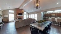 This single-story home showcases a thoughtfully designed open-concept layout that seamlessly connects the living, dining, and kitchen areas. The living room commands attention with its soaring vaulted ceiling accented by exposed white-painted beams, creating a sense of generous vertical space. A full-height brick fireplace serves as a dramatic focal point, complete with a white mantel and a mounted flat-screen television above. The room is bathed in natural light from multiple windows fitted with white shutters, enhancing the warm and welcoming atmosphere. Herringbone-patterned brick flooring runs throughout the main living area, adding a classic, textural character to the space. The dining area is positioned centrally, anchored by a rectangular marble-top table and black dining chairs, illuminated by a pair of striking geometric black metal pendant chandeliers. These fixtures echo one another and serve as elegant design statements. Adjacent to the dining space, the kitchen is visible through an open passthrough, revealing dark wood cabinetry and stainless steel appliances. Recessed lighting throughout the ceiling complements the natural light, ensuring the space remains bright and inviting at all hours.