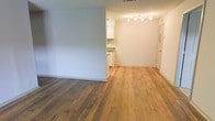 This single-floor residence offers a clean, open layout that maximizes the sense of space and functionality. The living area is anchored by warm-toned hardwood flooring that flows seamlessly throughout, lending a cohesive and inviting character to the interior. Neutral-painted walls in soft, light tones keep the space feeling bright and airy, complementing the natural warmth of the floors. The kitchen is partially open to the main living area, creating a connected feel between the spaces. It features white cabinetry, granite countertops, and a practical layout that makes efficient use of the available space. A track lighting fixture overhead ensures the kitchen area is well-lit and functional. The textured ceiling adds a subtle architectural detail consistent throughout the unit. A hallway leads to additional areas of the residence, with a door visible at the end providing clear separation between the living and private spaces. The overall ambiance is understated and clean, offering a well-maintained, move-in-ready environment with a timeless aesthetic that serves as a welcoming canvas for personalization.