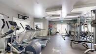 Located at 553 Settlers Landing Rd in Hampton, Virginia, this fitness center offers a well-equipped workout space on a single floor. The room features neutral gray walls, durable rubber-flooring, and bright overhead panel lighting that creates a clean, functional atmosphere throughout.

The cardio section is anchored by a row of Precor treadmills and elliptical machines, providing ample space for aerobic training. Opposite, a dedicated strength training area includes a cable machine, a weight bench with barbell setup, and a multi-function rack suited for a variety of resistance exercises. A medicine ball rack stocked with an assortment of weighted balls and a stationary bike round out the functional fitness options, catering to diverse workout styles.

Natural light filters in through multiple windows and a set of double glass doors, adding brightness and a sense of openness to the space. Full-length mirrors along one wall enhance the room's depth while allowing users to monitor their form. Additional conveniences include a wall-mounted water fountain, a sanitation station with cleaning supplies and paper towels, and a mounted television for entertainment. The space is also equipped with clearly marked emergency exits, ensuring safety throughout. Overall, this fitness center is a practical, well-organized facility designed to support a comprehensive range of exercise routines.