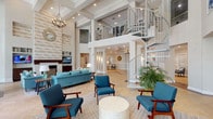 This expansive facility showcases an impressive architectural design centered around a grand common area with soaring double-height ceilings supported by structural columns. A striking white spiral staircase serves as a focal point, elegantly connecting the main level to an upper mezzanine overlook with decorative iron railings. An ornate chandelier adds a refined touch overhead.

The main lounge area features a coastal-inspired color scheme, with teal and blue upholstered seating arrangements set atop patterned area rugs over light travertine tile flooring. A fireplace with a decorative shiplap accent wall anchors one side of the room, complemented by built-in shelving and a mounted television.

Floor-to-ceiling glass walls and wide double-door entries flood the interior with abundant natural light while providing clear views of an outdoor courtyard. A secondary seating nook with built-in shelving and a mounted TV offers additional gathering space.

A well-equipped kitchen features rich, dark espresso cabinetry with glass-front uppers, granite countertops, a green tile backsplash, stainless steel appliances, and tile flooring. A transitional hallway with wainscoting, crown molding, and decorative accents connects the various functional areas of the facility, creating a cohesive and welcoming overall ambiance.