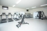 Located in Saint Clair, Michigan, this fitness facility is designed to accommodate a comprehensive range of exercise needs within a single-floor layout. The space is bright and clean, featuring white walls and light-toned flooring throughout, creating an open and welcoming atmosphere conducive to focused workouts.

The main workout area is generously sized, housing a lineup of cardio equipment that includes multiple treadmills and elliptical machines, all positioned near vertical-blind-covered windows that allow natural light to filter into the space. Overhead fluorescent lighting ensures consistent illumination throughout the room. Wall-mounted televisions are positioned for easy viewing during cardio sessions.

On the strength training side, a multi-station cable machine serves as the centerpiece, complemented by a dumbbell rack, weight benches, and resistance equipment. Exercise mats and foam rollers are also available, supporting flexibility and recovery work. Two wall-mounted split HVAC units provide climate control throughout the facility.

A dedicated utility area includes dual wall-mounted drinking fountains at accessible heights, a wall-mounted television, and seating. A corridor connects this area to additional rooms, suggesting supplementary spaces such as restrooms or changing areas. Safety features, including a fire extinguisher and exit signage, are also present.